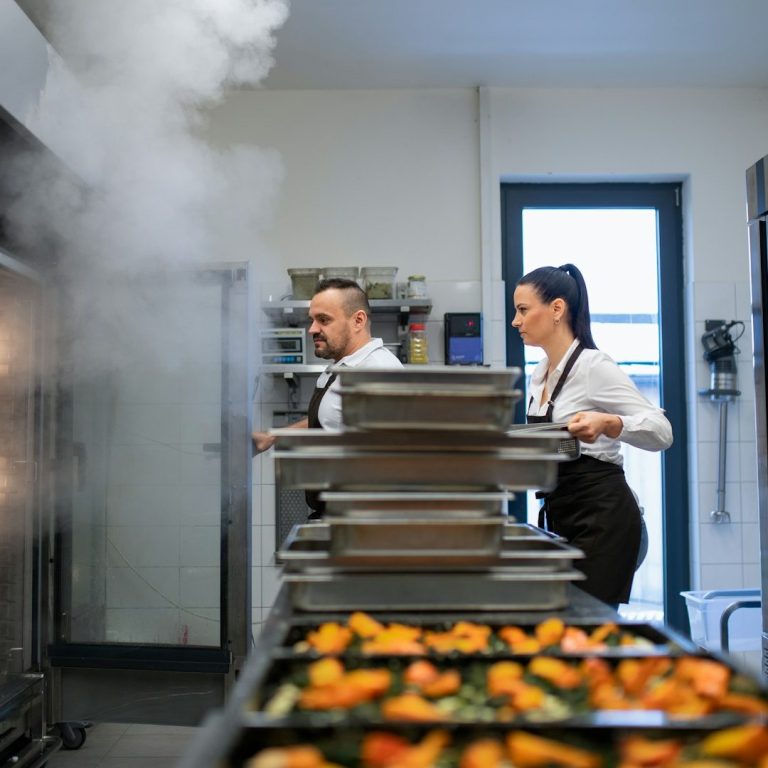 Küchenmitarbeiter bereiten Gerichte vor, dampfend mit Lebensmitteln auf Blechen.
